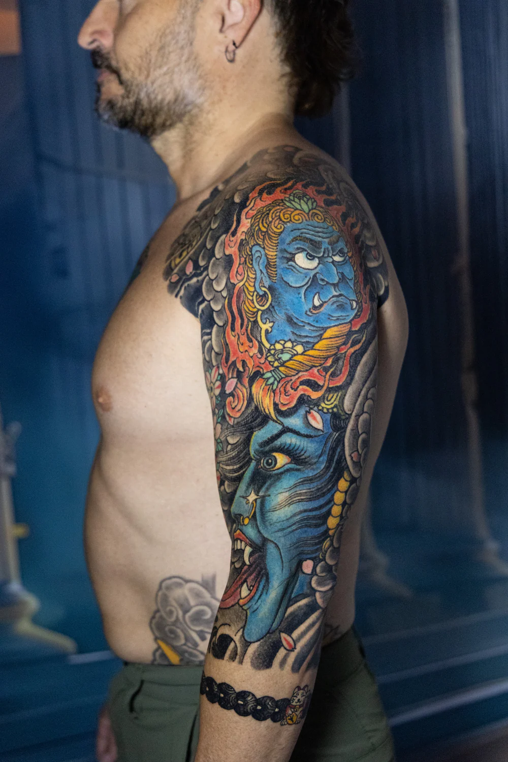 Tatuaje de brazo para hombre: media manga japonesa a color con Fudō Myōō entre llamas, cara azul, nubes y flores de cerezo